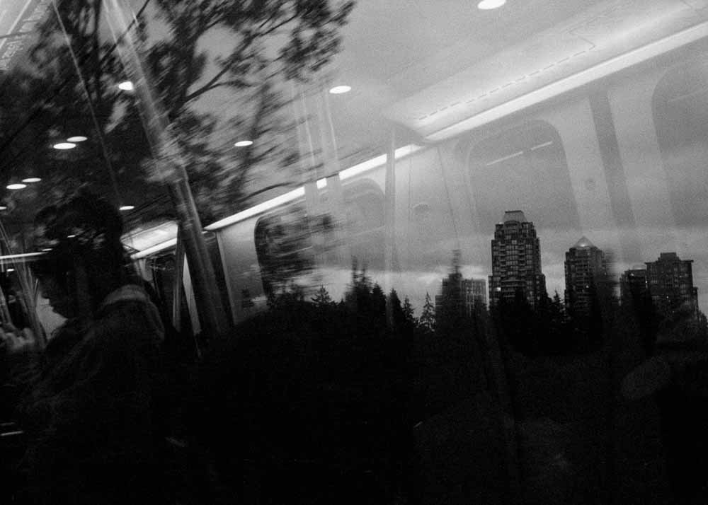 Twilight. Silhouettes of buildings and trees on the horizon. A large, blurry tree blends with a reflected passenger.