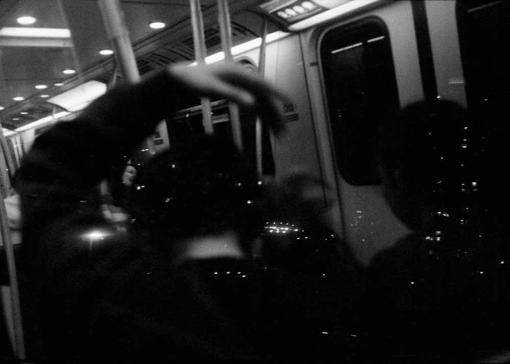 Night. Specks of light. Reflected: two seated passengers, one with their arm arced above their head, reaching.
