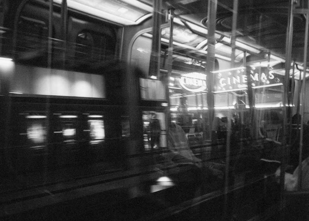 Night. A sign for a cinema, and some blurry backlit posters. Reflected: passengers, their heads obscured by the exterior lights.