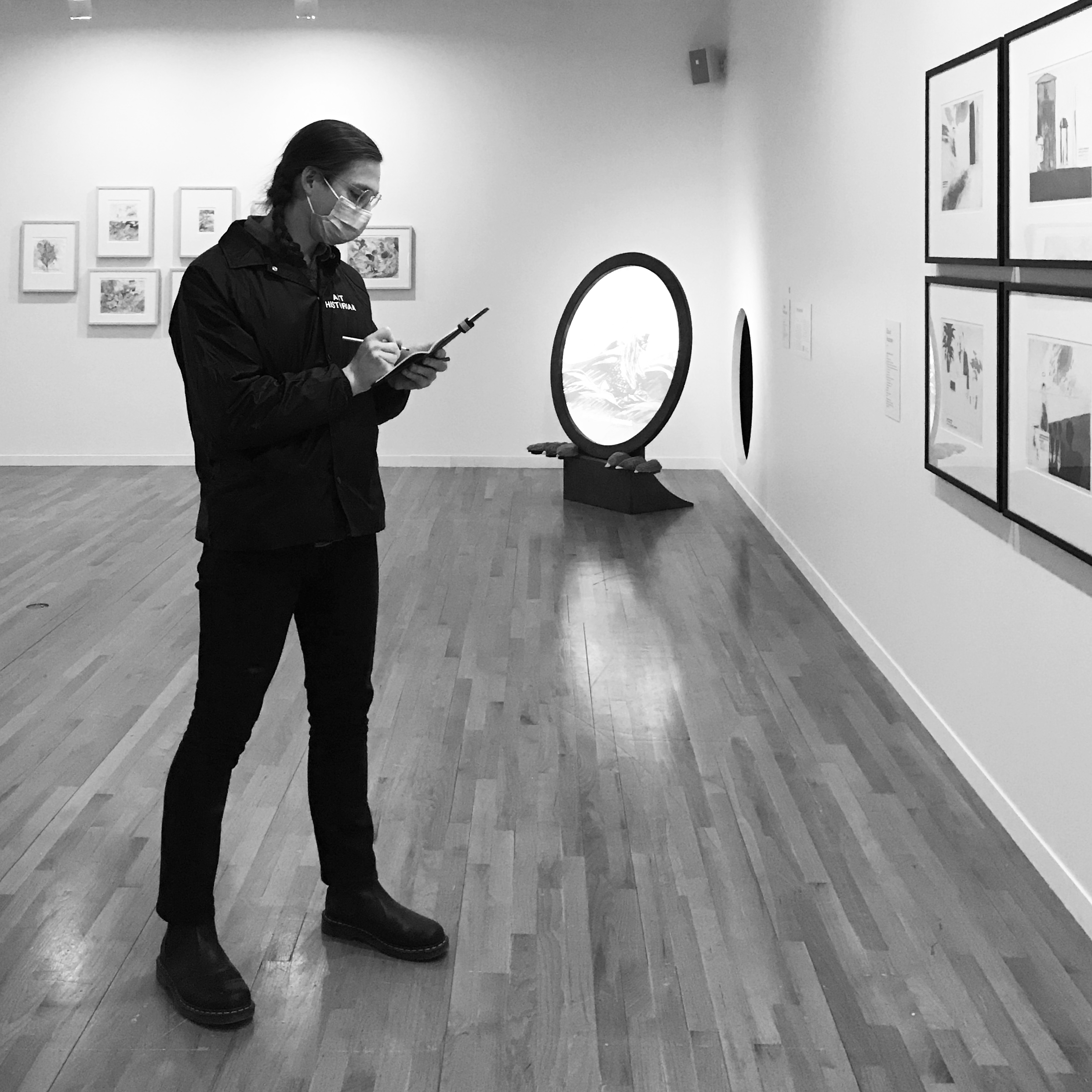 From the side: me wearing the jacket and writing in a police-style notepad, while facing a series of drawings on a gallery wall.