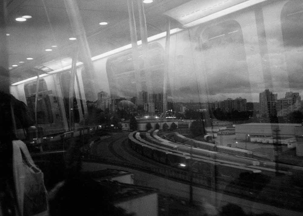Day. An industrial area, curving train lines, silhouettes of buildings. Reflected: windowframes, handholds, a tote bag.