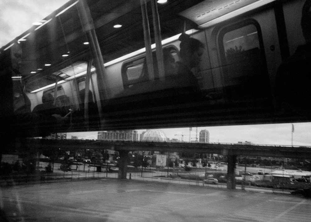 Day. An overpass. Below that, Science World, another overpass, parking. Reflected: passengers, one with hair in a bun.