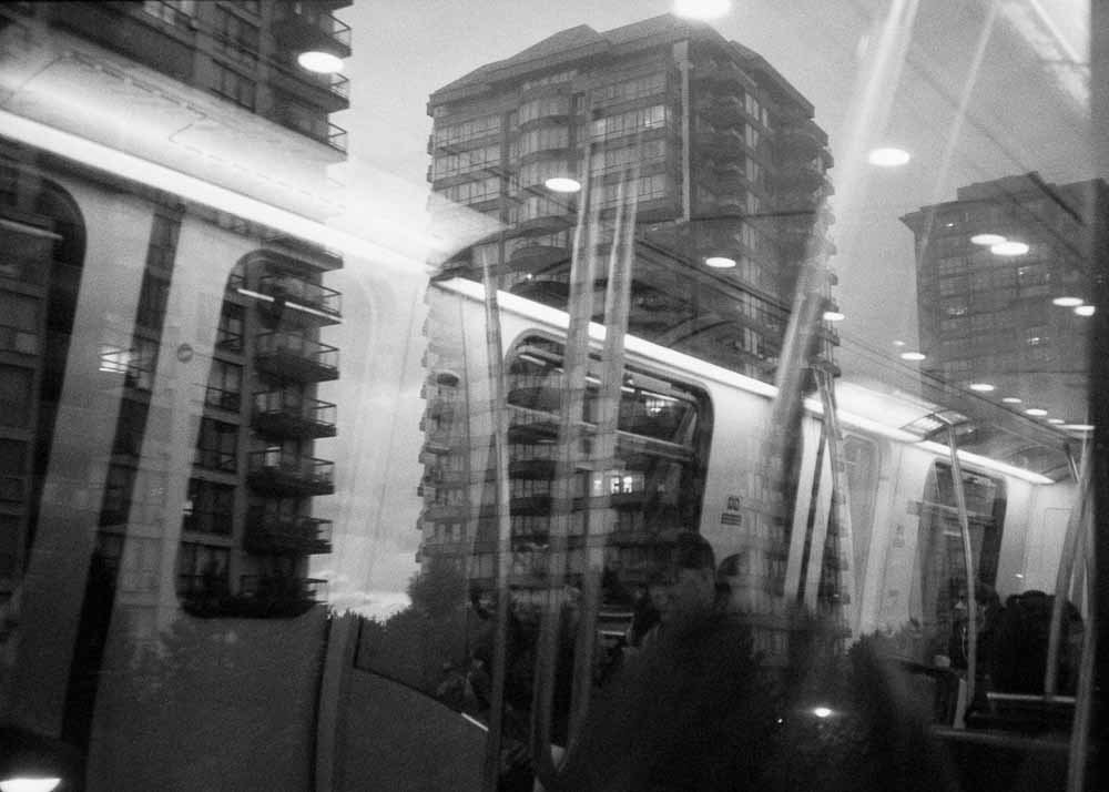 Day. Apartments. Reflected: bright white panels between train windows, handholds, seated passengers.