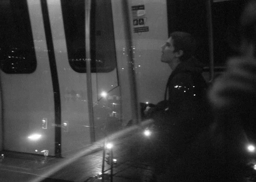 Night. Traffic lights below. Reflected: a man seated by the doors, with his mouth slightly open.