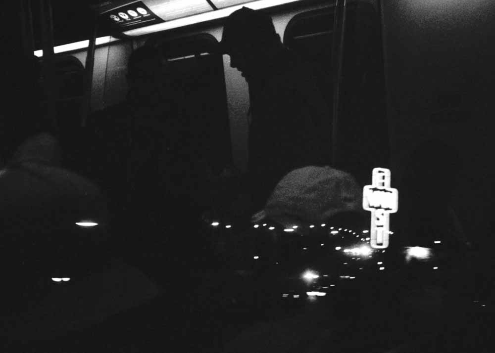 Night. The East Van Cross. Scattered streetlights. Reflected: passengers with caps, one white, one in black silhouette.