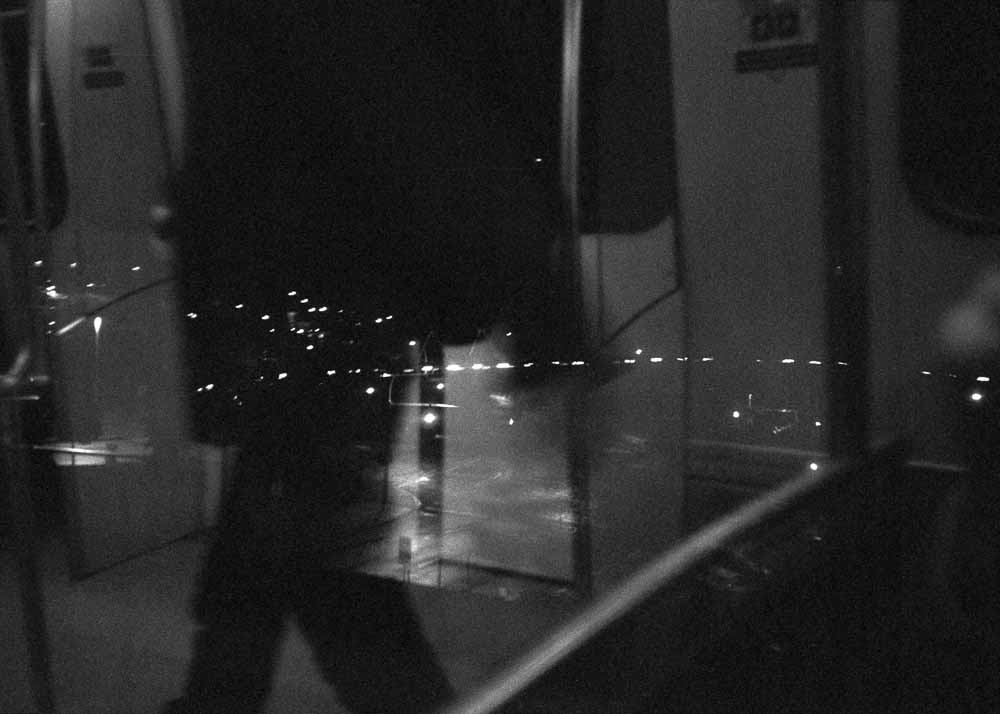 Night. An arcing, dotted line of lights, leading to a scattered cluster. Reflected: a passenger walking, in silhouette.