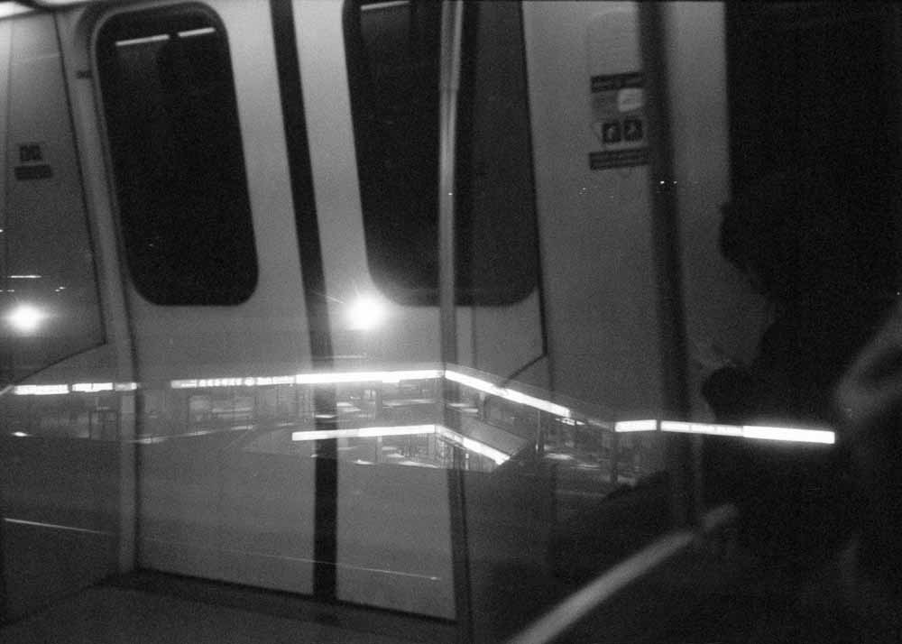Night. Angled stripes of light from a mall, and two round bursts of light from lampposts. Reflected: the train doors.