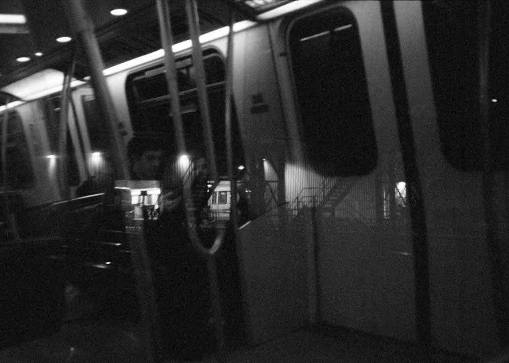 Night. The metal staircases and corrugated walls of a loading bay. Reflected: train furnishings, two passengers.