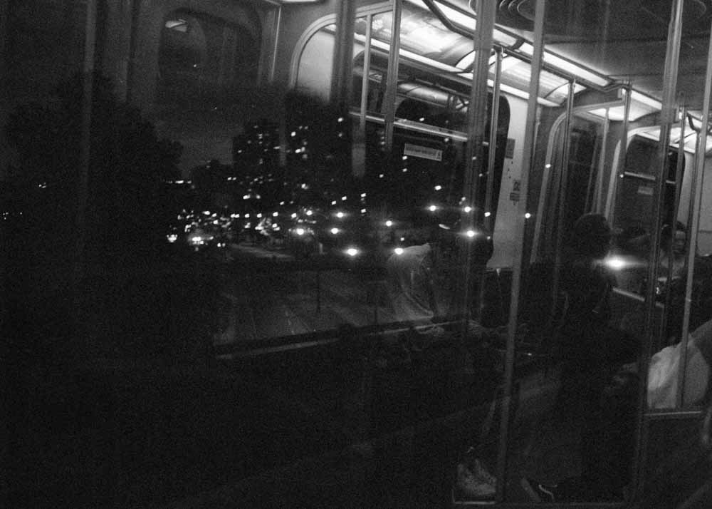 Night. Many scattered bright spots from streetlights. Reflected: bright backlit ads, handholds, and a few passengers.