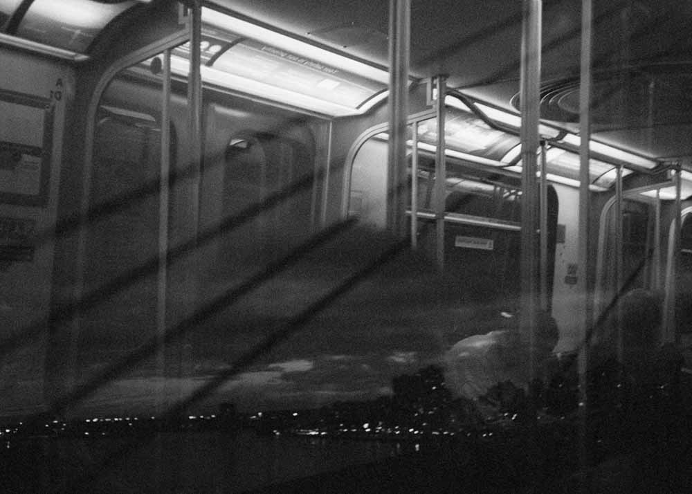 Night. The horizon, and five dark lines diagonal lines. Reflected: a passenger with a hat and hunched shoulders.