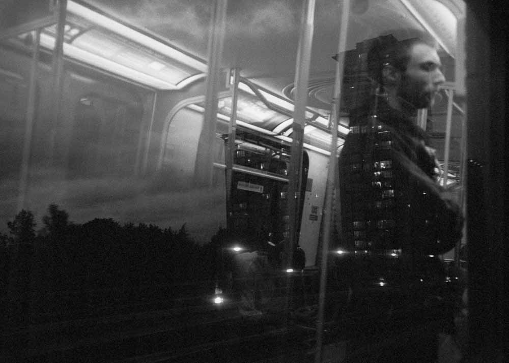 Twilight. Trees, streetlights, apartment windows.  Reflected: a profile view of a young man standing with arms crossed.