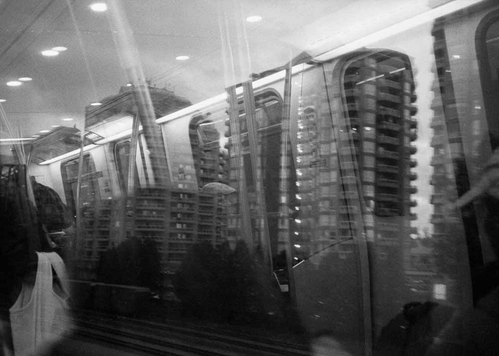 Day. Apartments. Balconies make a dark pattern against a light sky. Reflected: partial, blurry passengers, and a tote bag.