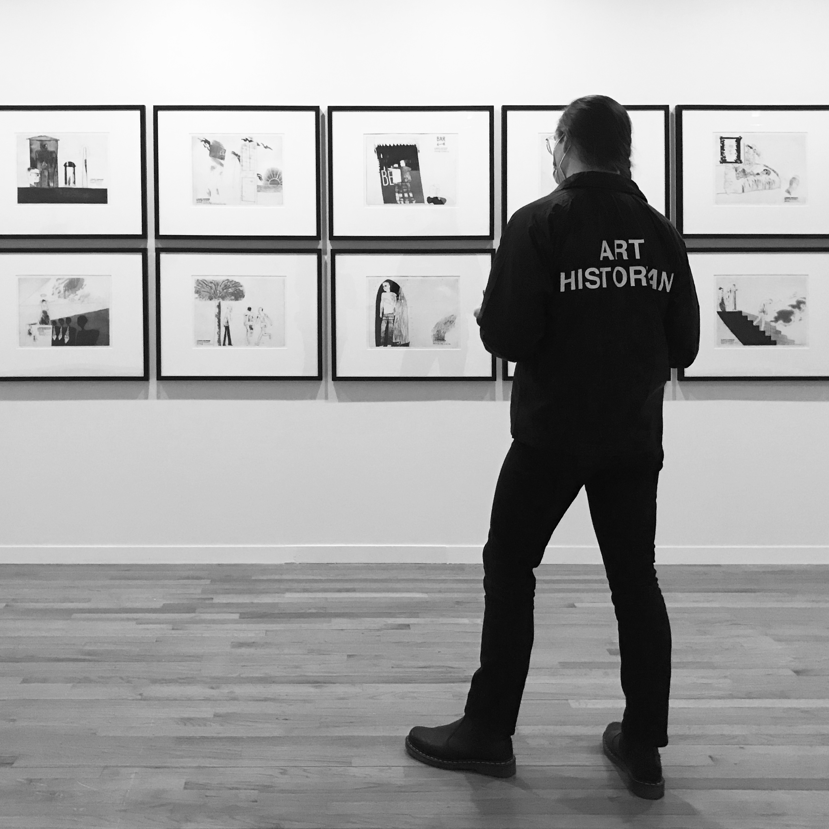 From behind: me wearing the jacket, with 'art historian' visible on the back. I am looking at a series of framed drawings on a gallery wall, and wearing black pants and black boots that match the jacket.